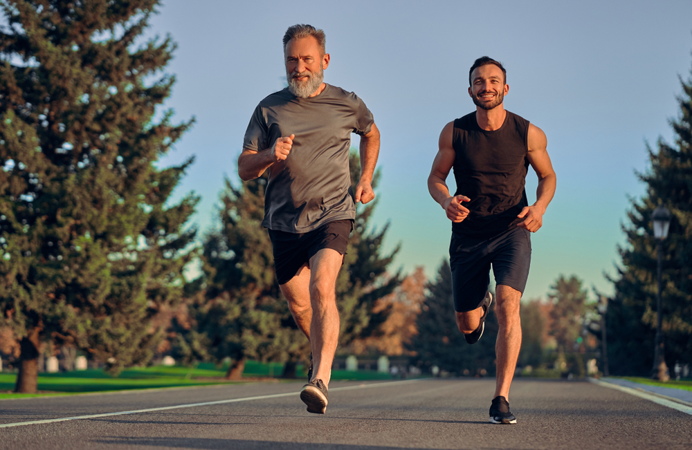 Smiling, athletic couple jogging in a sunny, forested area, enjoying an energetic workout in nature - Butler Spine & Sport The happy old and young sportsmen running on the road - Sports Rehab in Overland Park