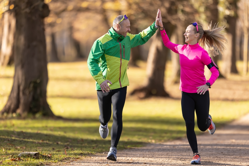 Smiling, athletic couple jogging in a sunny, forested area, enjoying an energetic workout in nature - Butler Spine & Sport